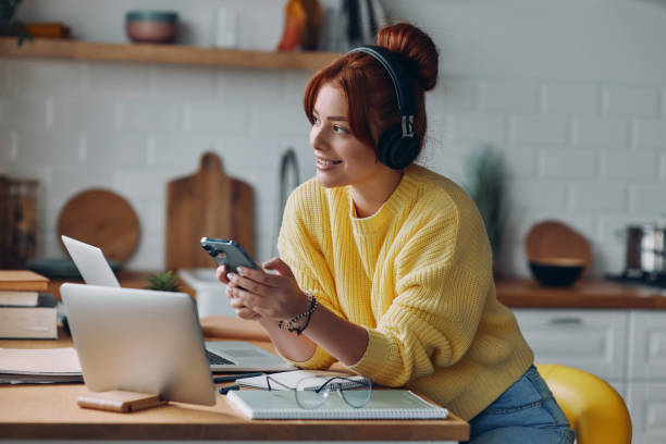 istock Joyful young woman in headphones using technologies while sitting at the kitchen counter 1437699417