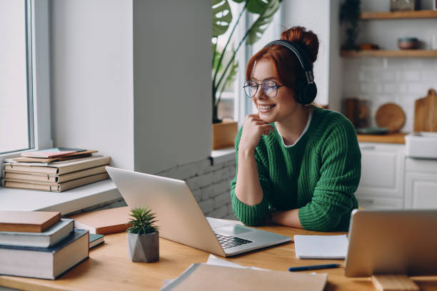Beautiful young woman in headphones looking at laptop and smiling while working from home stock photo