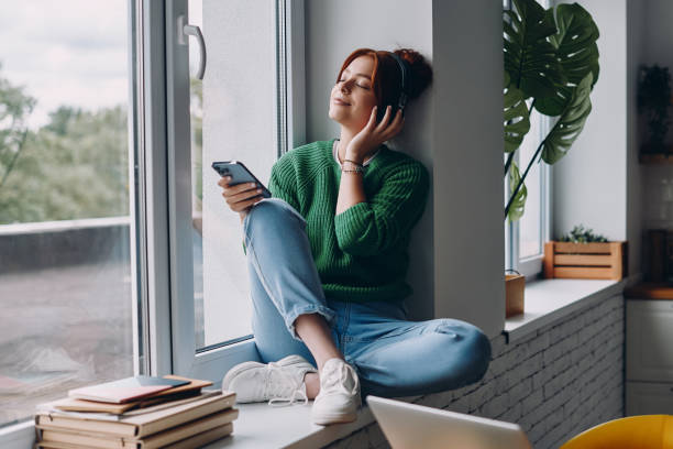 zamyślona kobieta w słuchawkach trzymająca smartfon, siedząca na parapecie w domu - window sill audio zdjęcia i obrazy z banku zdjęć