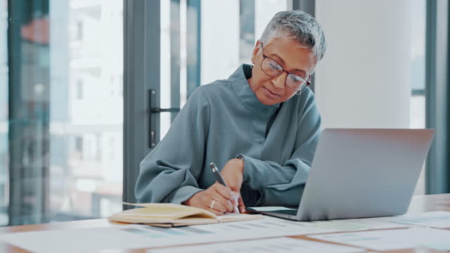Mature businesswoman writing and using a laptop in an office. Entrepreneur writing in a journal while taking notes for a business plan at a corporate startup company. A secretary scheduling meetings