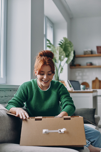 Excited young woman unpacking box while sitting on the couch at home