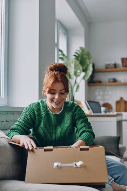 Excited young woman unpacking box while sitting on the couch at home stock photo