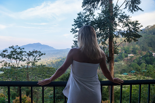 Rear view tourist young woman relaxing on balcony and looking mountain landscape at countryside Sri Lanka wearing towel in morning. Vacation, travel, journey, trip and relax concept. Copy text space