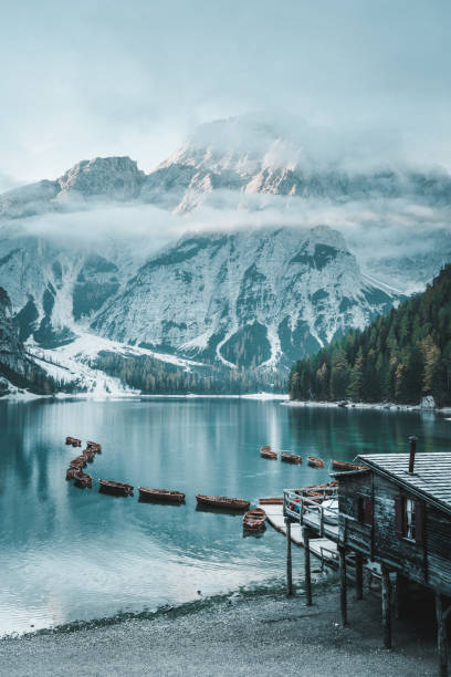 Vertical shot of mountain lake with empty wooden row boats on a autumn morning, some clouds, fog close to mountains in background and larch trees along the beach