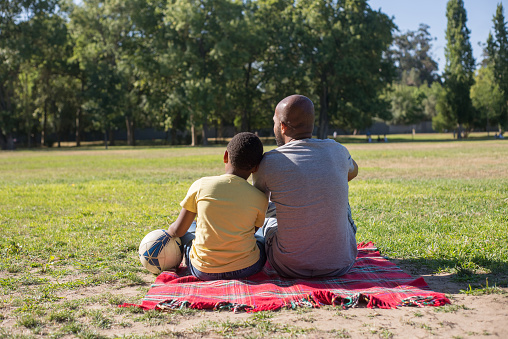Back view of dad and son sitting close on ground in summer. African American man resting on blanket and boy leaning with his head on fathers shoulder. Fatherhood and having rest together concept