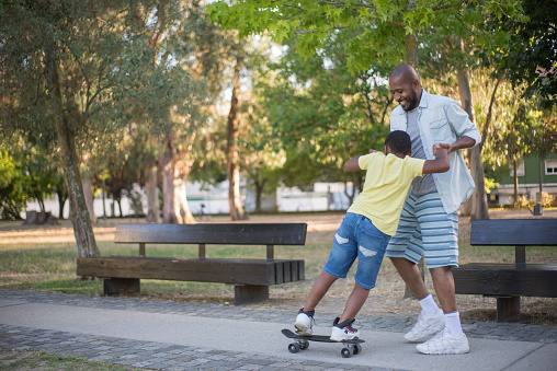 Portrait of father teaching his son to skateboard. Smiling Black man standing holding boy hands helping not to fall down and kid trying to balance. Kid healthy growing-up and happy childhood concept
