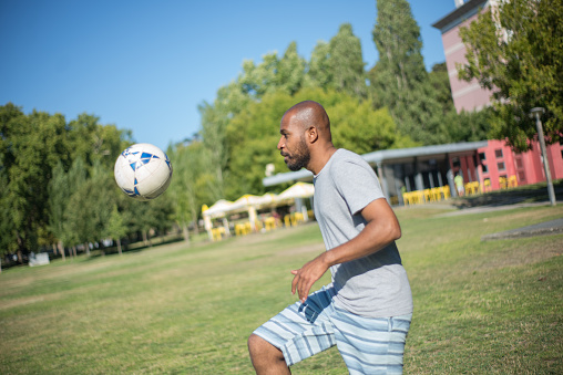 Side view of sporty African American man playing ball in park. Young man concentrated standing on grassy ground kicking ball. Leisure, active rest and healthy lifestyle concept