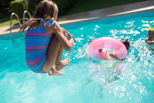 Back view of girl in swimsuit jumping into swimming pool Back view of girl in swimsuit jumping into swimming pool. Little girl in swimsuit falling down into water holding her knees looking at other children in pool. Summer vacation, happy childhood concept young hot stock pictures, royalty-free photos & images