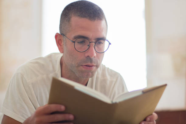 Focused man in eyeglasses reading book at home in morning. Close-up shot of cheerful man reading hardcover book with interest, having rest on weekend. Hobby, education concept