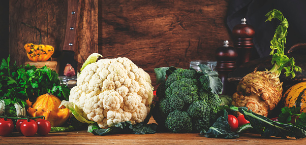 Autumn food background with organic farm vegetables: cauliflower, broccoli, root celery, pumpkin, herbs and spices on rustic wood kitchen table with cast iron pan, spice grinders, cutting board. Banner