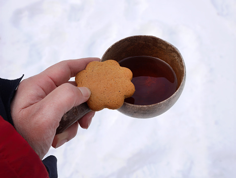 High angle view a hand holding biscuit cookie with warm drink in a wooden cup outside under white snow covered, happy holidays