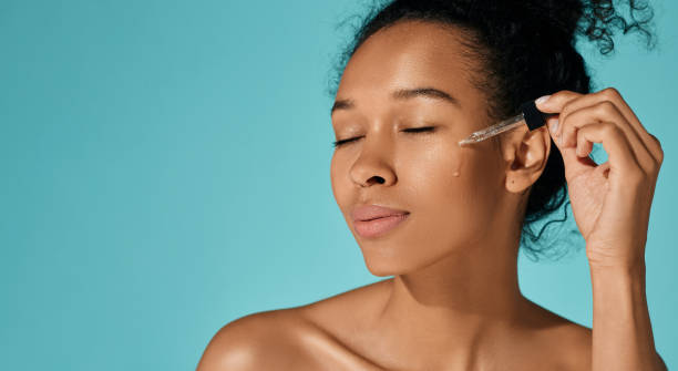 Facial skin care with essential oil and serum. Happy African woman applying essential oil on her face for hydrated skin with pipette dropper, isolated on blue background Facial skin care with essential oil and serum. Happy African woman applying essential oil on her face for hydrated skin with pipette dropper, isolated on blue background face serum stock pictures, royalty-free photos & images