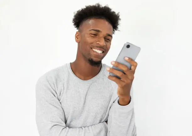 Portrait of happy African American man using social network on mobile phone. Young bearded guy wearing white sweater reading message on smartphone and smiling. Mobile technology concept