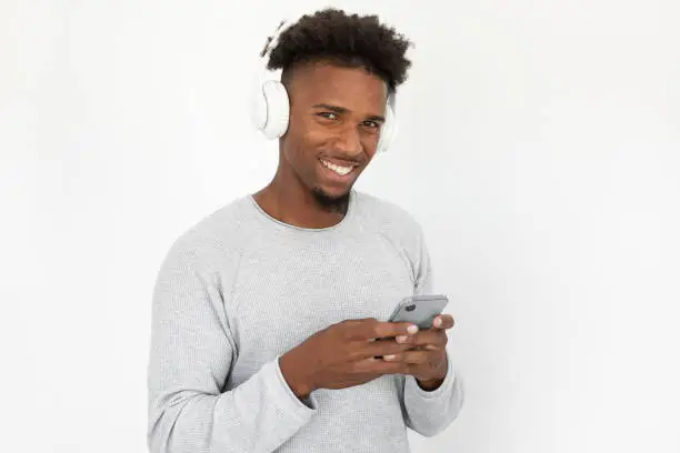 Portrait of happy young man in headphones using smartphone. African American bearded guy wearing white sweater texting message, looking at camera and smiling. Hobby and music concept
