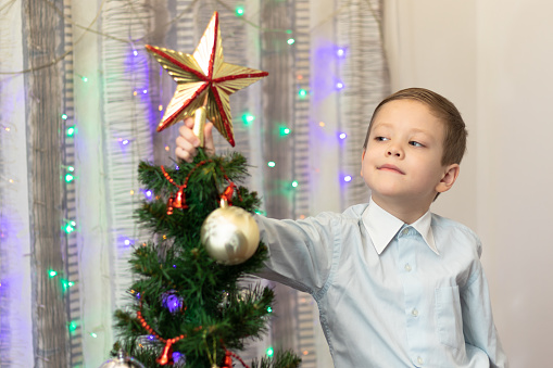 A seven-year-old boy in a shirt decorates a Christmas tree with toys on New Year's Eve at home against a background of garlands. Selective focus. Portrait