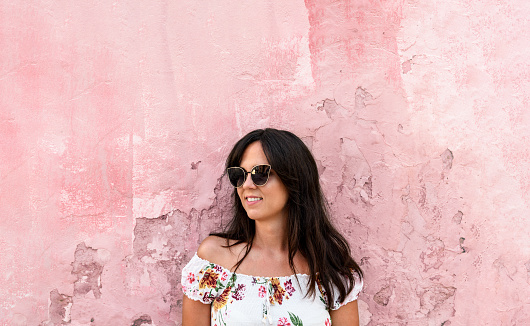 A closeup of a female wearing a floral dress and butterfly sunglasses standing  on the pink wall