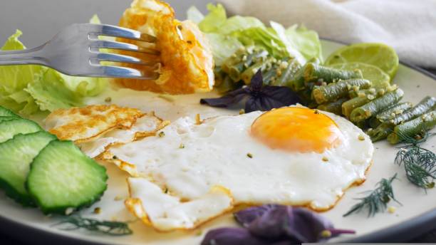 istock Close-up of a fork taking a fried egg yolk from a plate On a white plate, side view, a portion of fried eggs, fresh lettuce leaves, chopped cucumber and lemon slices, asparagus and greens Healthy food 1437725236