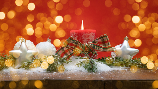 Christmas backgrounds: white burning Christmas candle shot on rustic wooden table. The composition is at the left of an horizontal frame leaving useful copy space for text and/or logo at the right.\nString light and Christmas decoration are out of focus at background. Predominant colors are red and brown. High resolution 42Mp studio digital capture taken with Sony A7rII and Sony FE 90mm f2.8 macro G OSS lens