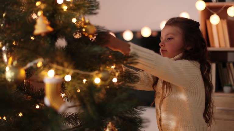 Little adorable girl in knitted sweater is hanging decoration on Christmas tree with light.