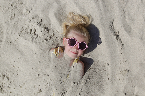 Cute happy little girl buried in the sand on the beach.