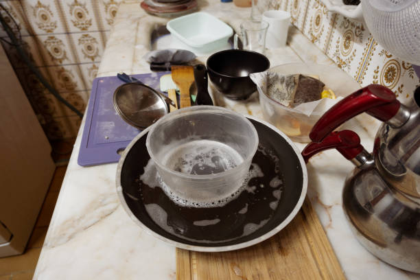An assortment of stainless steel cookware including large mixing bowls, saucepans, and stock pots sits on a white quartz countertop in a contemporary kitchen. Nearby, a double stainless steel sink holds a yellow sponge and dish cloth, with bottles of olive oil, vinegar, and a green cleaning solution placed adjacent. Black wooden cabinets with silver handles line the base, and a window allows natural daylight to illuminate the scene. The image captures a cluttered yet functional domestic setting indicative of meal preparation or post-cooking cleanup. Ideal for commercial use in recipe illustrations, home cooking tutorials, kitchen organization guides, appliance advertising, and lifestyle content for media outlets focusing on everyday homemaking and culinary activities.