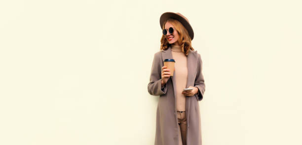 Portrait of stylish beautiful young woman model with cup of coffee wearing brown round hat and coat on beige background stock photo