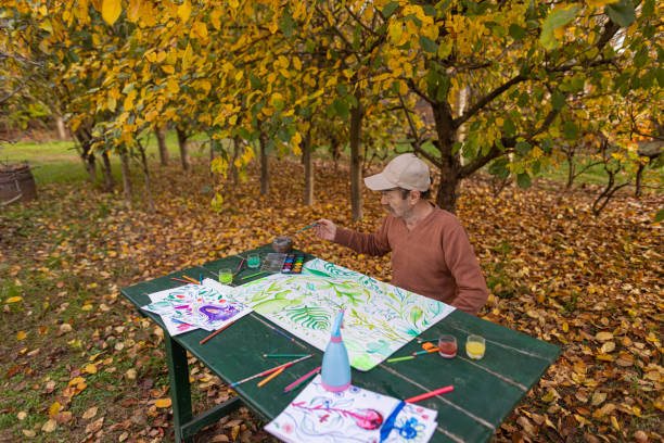 A shot of the mature man with a mustache, eyeglasses, and a cap sitting in the garden full of trees and falling leaves and painting with watercolors.