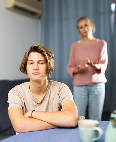 Frustrated teenage boy sitting at the table at kitchen after conflict with his mother