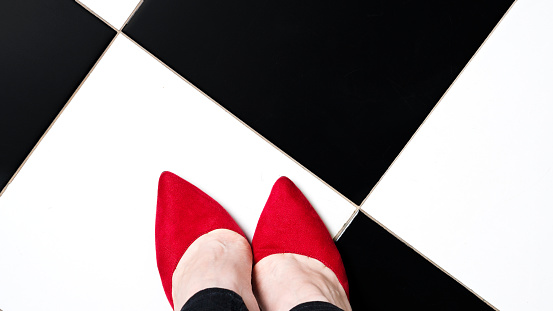 Selfie feet wears classic shoes court shoe on chequered floor background. Image legs in red velvet shoes pumps on black and white floor. Top view
