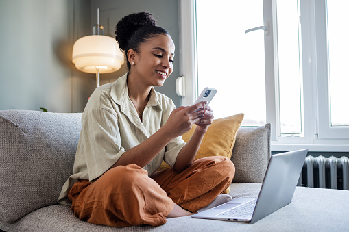 Young African American woman sitting comfortable on the couch and smiling while using mobile phone. A laptop is on the couch, too.