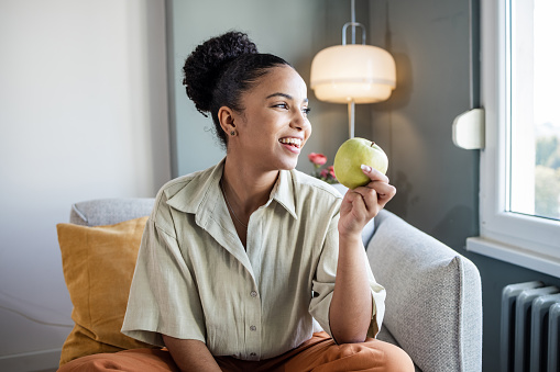 Smiling African American woman sitting on the couch and eating an apple. Healthy snack.