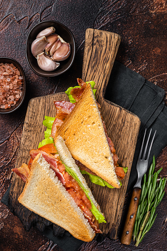 Classic BLT sandwich with bacon, tomato and lettuce on wooden board with herbs. Dark background. Top view.