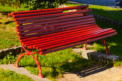 Wooden red park bench, sidewalk and garden path, shadows at dusk, green background. Vilagarcía de Arousa, Rías Baixas, Pontevedra province,  Galicia, Spain.