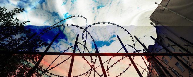 Jail building behind barbed wire against russian flag over cloudy sky