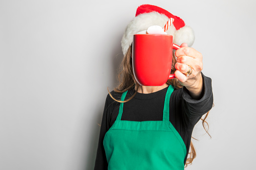 A barista in a Santa hat holding out a holiday hot drink that is covering her face.
