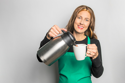 A middle aged barista in a green apron smiling as she pours a cup of fresh coffee, isolated on a white background.