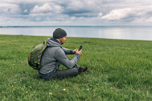 Tourist sitting on the green grass, holding his phone enjoing beautiful blue sea and claudy, moody sky. Horizontal photo.