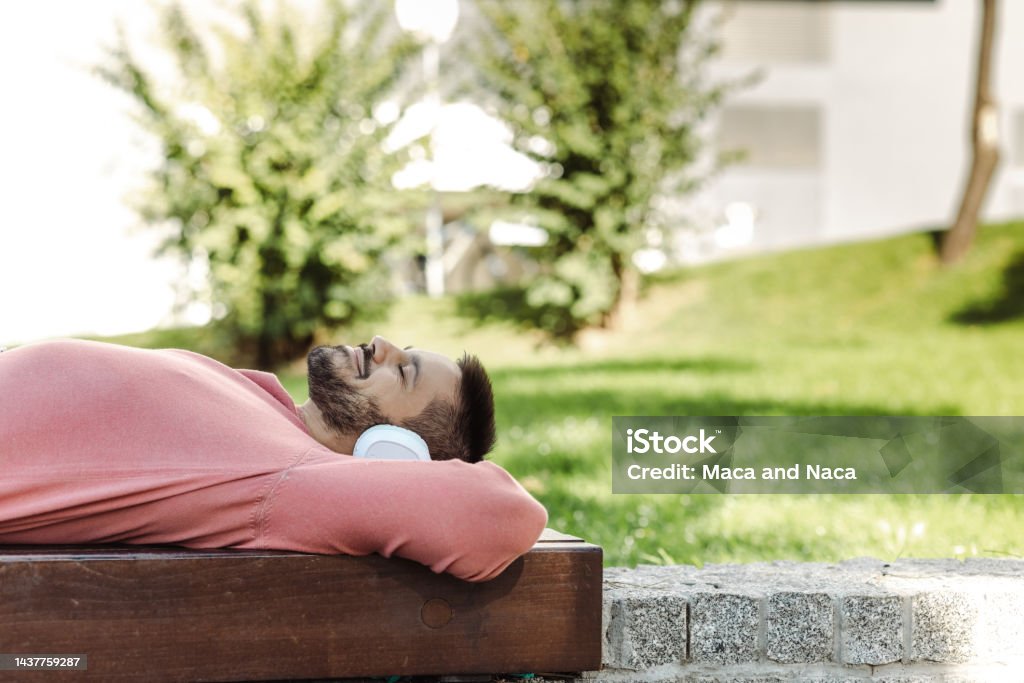 Lazy day at the city park Side view photo of a smiling man lying on a bench and listening to the music 25-29 Years Stock Photo