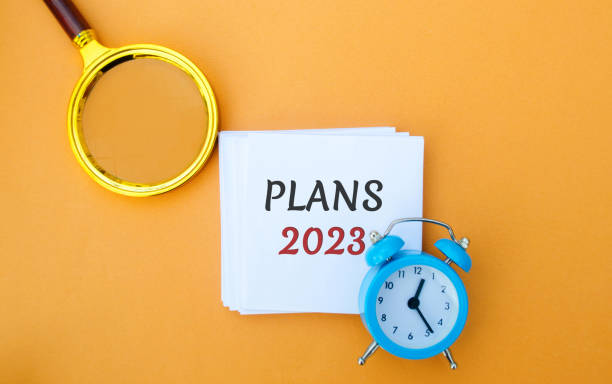 istock Note with words Plans 2023. Setting goals and planning targets for the next year. Motivation, vision. Business and finance concept. Magnifying glass and alarm clock 1437759916