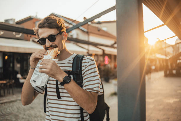 an adult fashionable man is walking in the city and eating a sandwich - lanche da tarde imagens e fotografias de stock