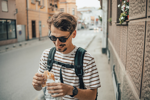 One man, adult fashionable man in brown white striped t-shirt walking in the city and eating a sandwich, concept of fast food and urban lifestyle in the city