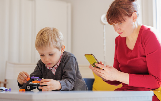 Toddler playing with toy truck on the living room table, his mother using smart phone and visiting web stores, online shopping consumerism