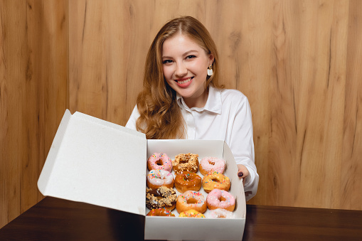 Happy young woman tasting new donut and Doughnut, enjoying sweet food in box. Concept with unhealthy eating and bakery indoors, person is holding and showing dessert in cafe or home