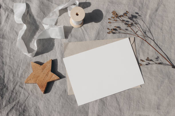 Christmas stationery. Blank greeting card, invitation mockup. Wooden star ornament, craft envelope and dry grass in sunlight. Beige linen tablecloth. White ribbon. Winter wedding flat lay, top stock photo