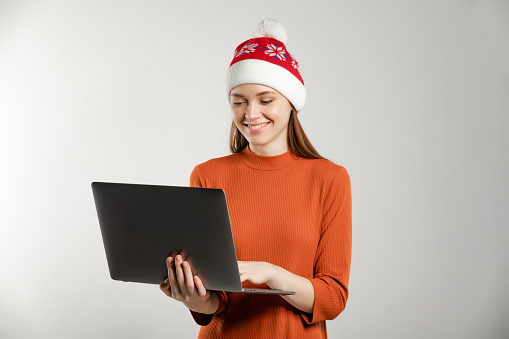 Christmas Santa hat concept. Portrait of young lady with at the studio with copy space and white background. Woman with emotions posing and gesturing. Photo with laptop