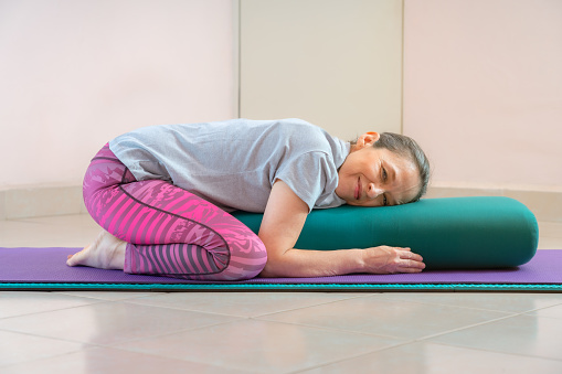 Shot of energetic mature woman making exercising and practicing restorative yoga on mat and bolster at home because of social distancing, wearing sports clothes.