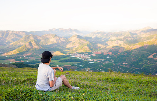 Man sitting on grass looking at landscape with valley and mountains in background. Young man sitting quietly and enjoying the moment outdoors.