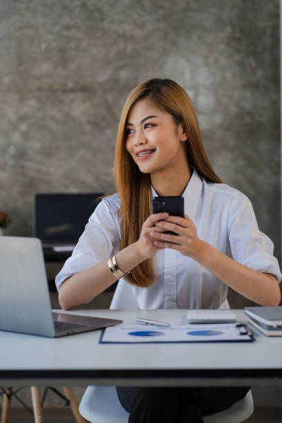 istock Asian female accountant holding smartphone working on laptop analyzing financial account data with graph document on table. 1437805209