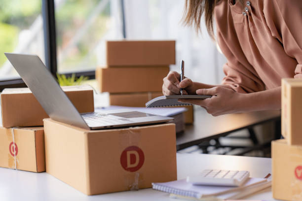 istock Smiling Asian woman looking at camera and using laptop to check package delivery information before sending it to customer. Small sme business entrepreneur. Online selling concept. 1437805472