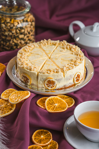 High angle studio shot of a round, home made cake arranged with dried orange slices, a cup of tea and a tea pot,  nuts in a jar in the background, all on purple fabric. Full length vertical  image.